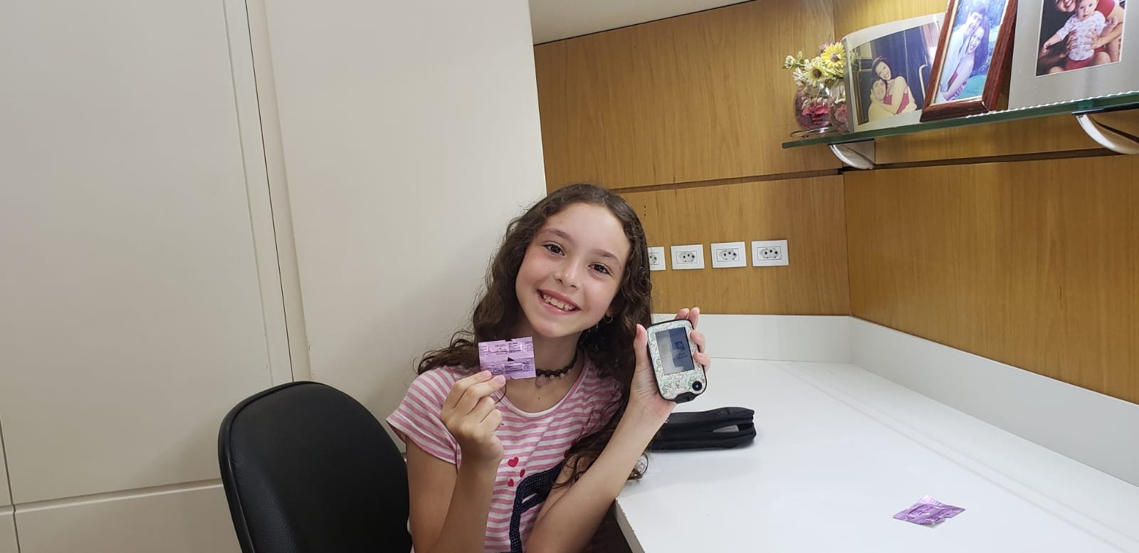 Livia, criança com diabetes tipo 1, sorrindo, com o estojo do glicosímetro aberto e as fitas na mão