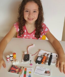 Livia, criança com diabetes tipo 1, à mesa com o kit do tratamento: canetas de insulina, glicosímetro, fitas, lancetador e frascos