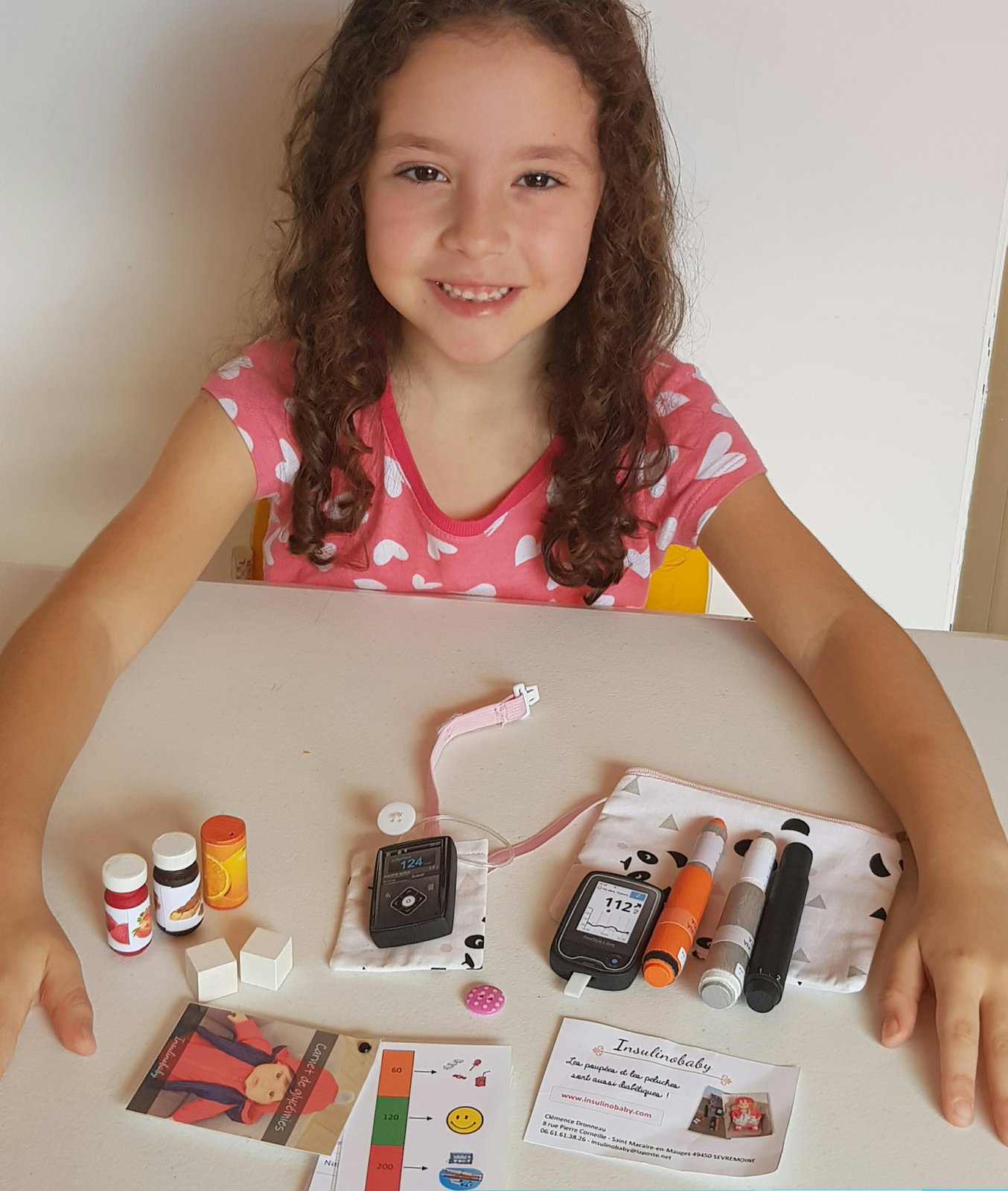 Livia, criança com diabetes tipo 1, à mesa com o kit do tratamento: canetas de insulina, glicosímetro, fitas, lancetador e frascos