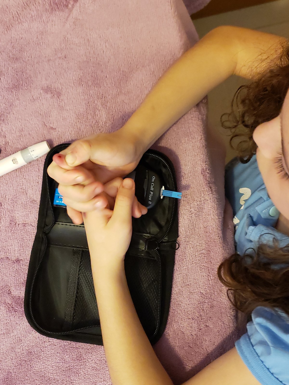 Livia, criança com diabetes tipo 1, deitada na cama medindo a glicemia na ponta do dedo com o kit aberto ao lado